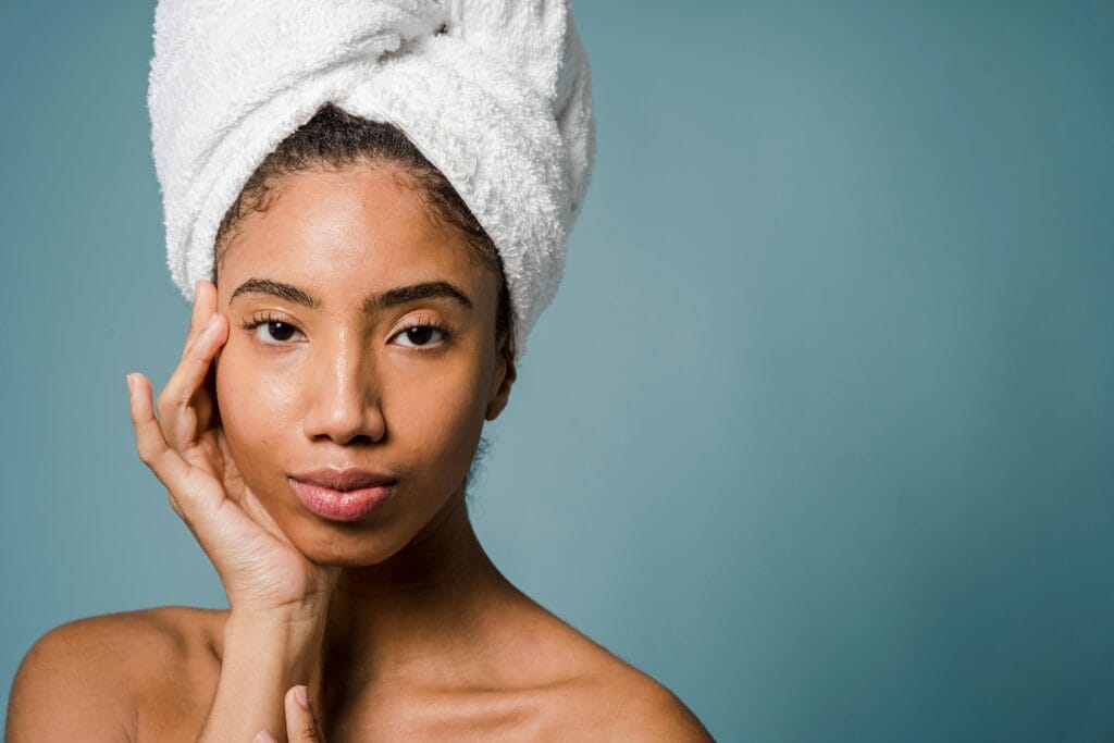 Almond Oil Tranquil young black female with pure skin and white towel on head touching face and looking at camera after spa procedures against blue background